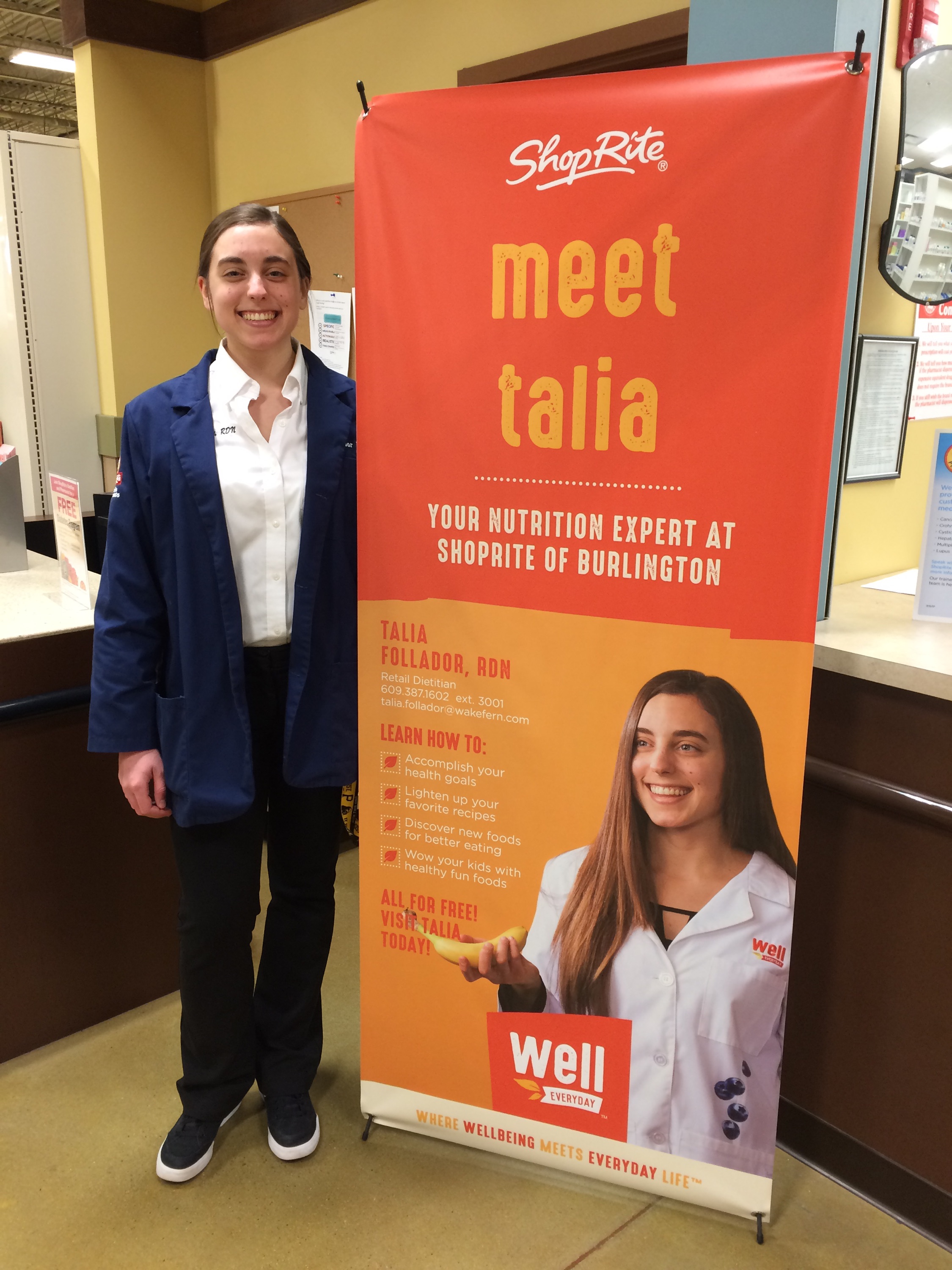 Girl standing next to large poster of herself.  Poster is orange and yellow. Poster has a picture of the girl holding a banana and smiling. Poster says: "Meet Talia. Your nutrition expert at ShopRite of Burlington."