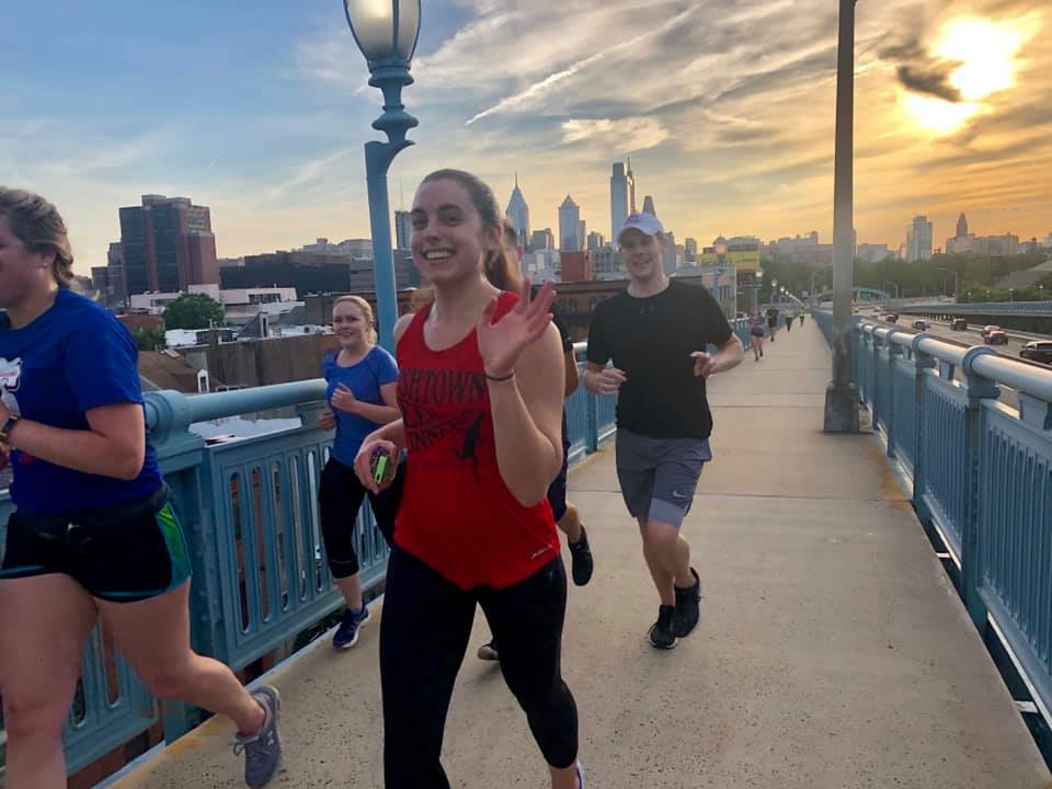 Girl running with a group of friends with the city sky line and sunset behind them. Girl is waving at the camera.